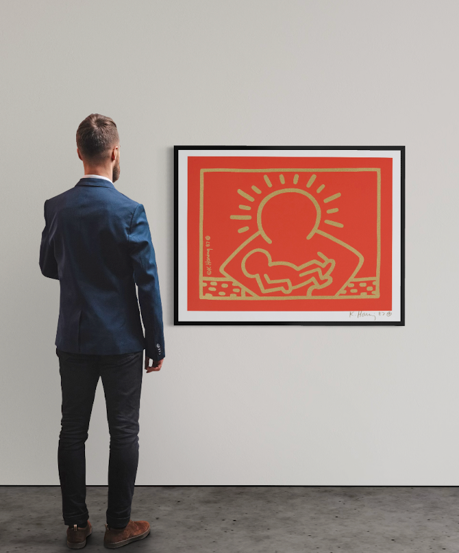<img alt="Installation view of Keith Haring's A Very Special Christmas, framed work displayed on a white gallery wall, with a visitor standing with their back to the camera contemplating the piece." />